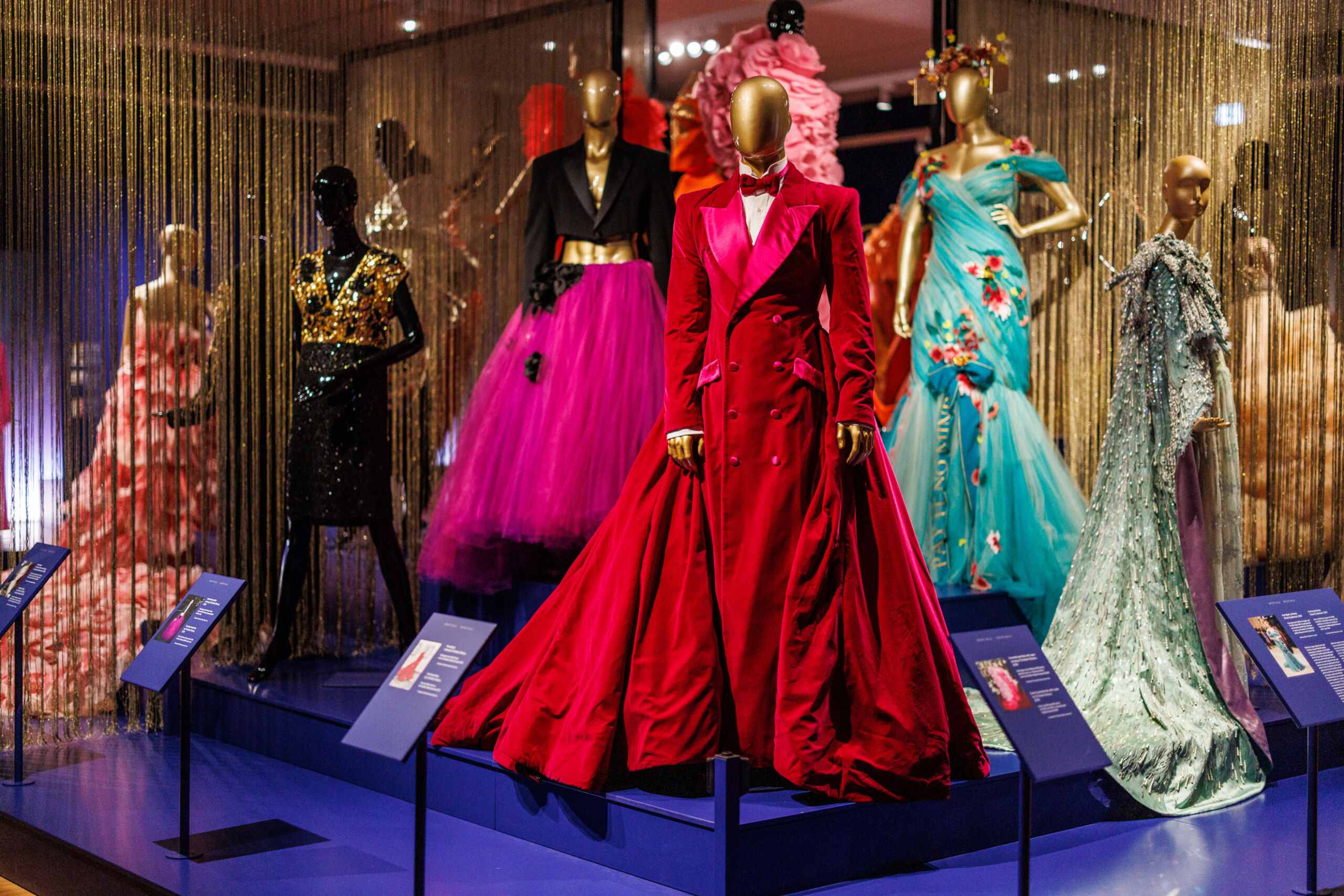 Tentoonstellingsruimte met geklede etalagepoppen in extravagante, kleurrijke couturejurken en kostuums. Centraal staat een gouden mannequin in een felrode, lange manteljurk met een wijde sleep. Daaromheen staan andere poppen in opvallende ontwerpen, zoals een turquoise jurk met bloemen, een roze tulejurk en een zwarte glitterjurk met gouden details. De setting is luxe, met gouden gordijnstrengen en blauwe informatiebordjes voor elke outfit.